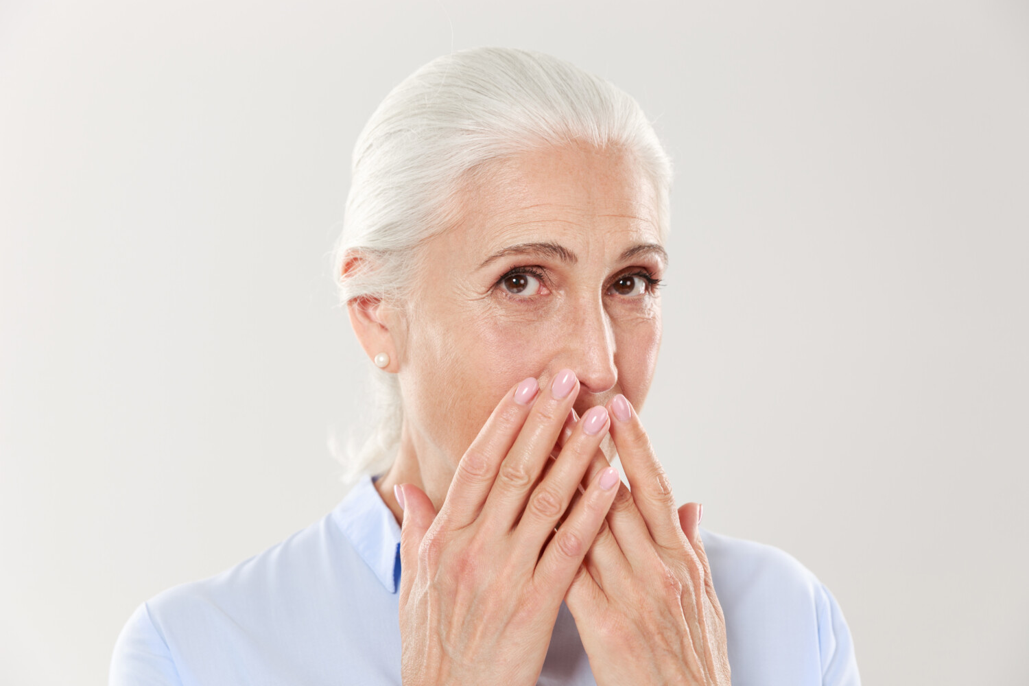 Close-up portrait of charming old lady, covering her mouth with hands, looking at camera, isolated over white background