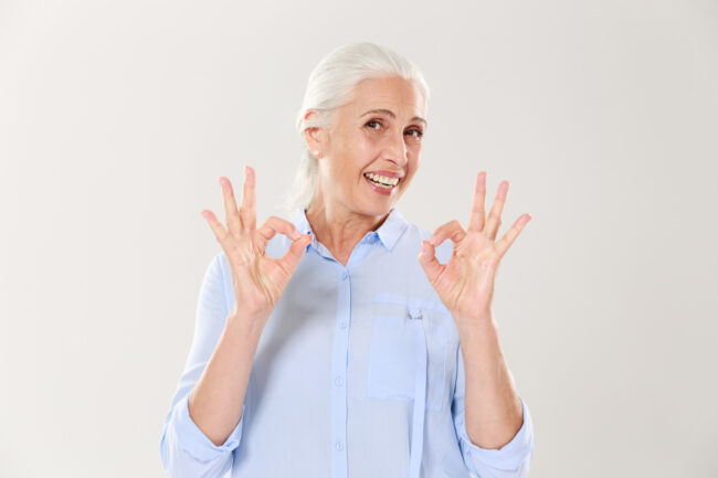 Portrait of smiling senior woman in blue shirt showing OK gesture, isolated on white background